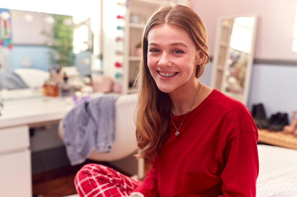 Portrait Of Teenage Girl Wearing Orthodontic Braces Sitting On Bed At Home